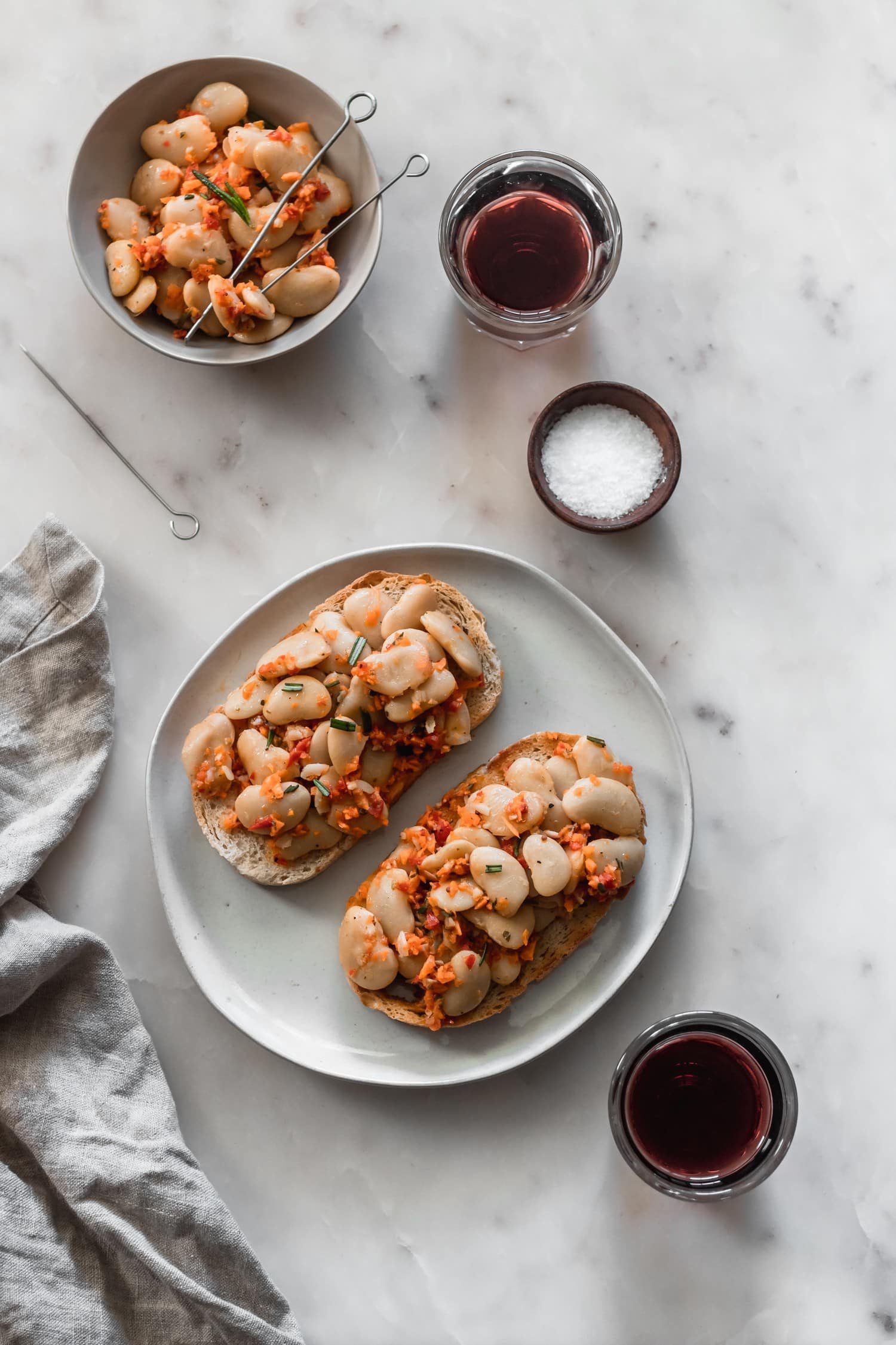 A white plate with toast topped with bean, carrot, and pepper salad. The plate is on a white marble board next to a beige linen, surrounded by wine glasses and a small bowl of salt.