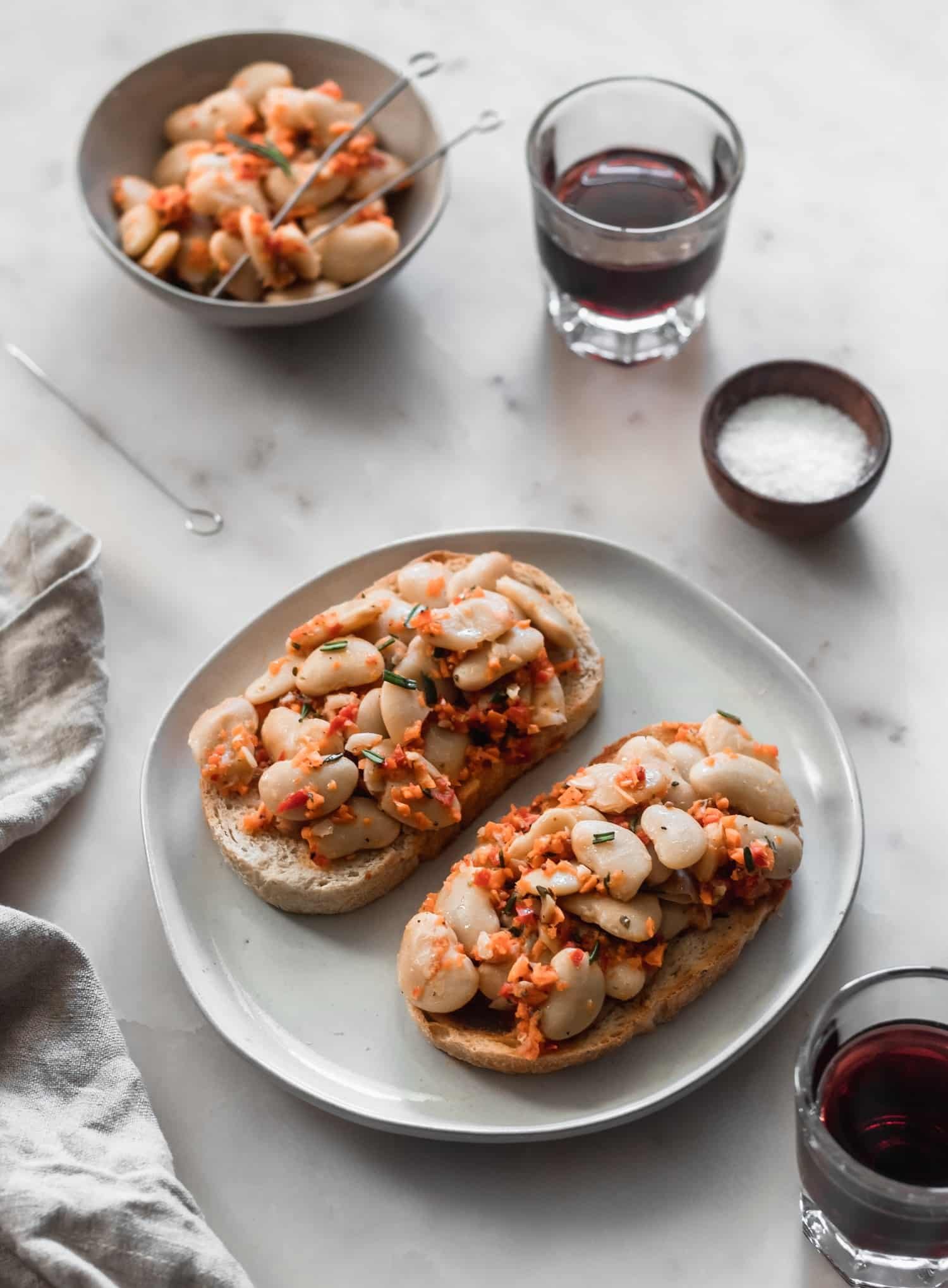 A 45 degree shot of a plate of limas, vegetables, and toast on a white marble backdrop surrounded by wine glasses, a beige linen, and a small beige bowl of limas.