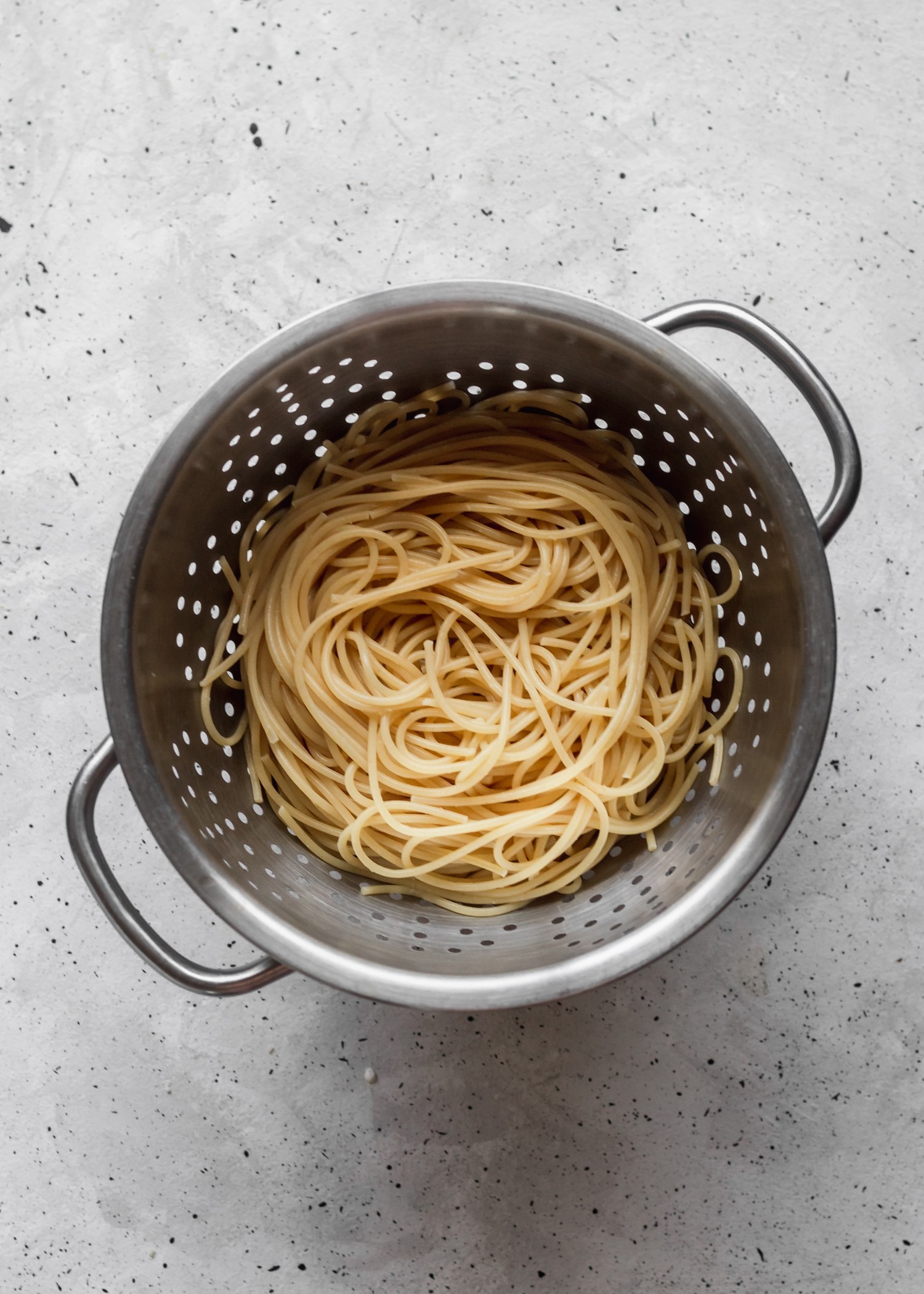 A bird's eye photo of a pasta strainer filled with spaghetti on a grey speckled table.