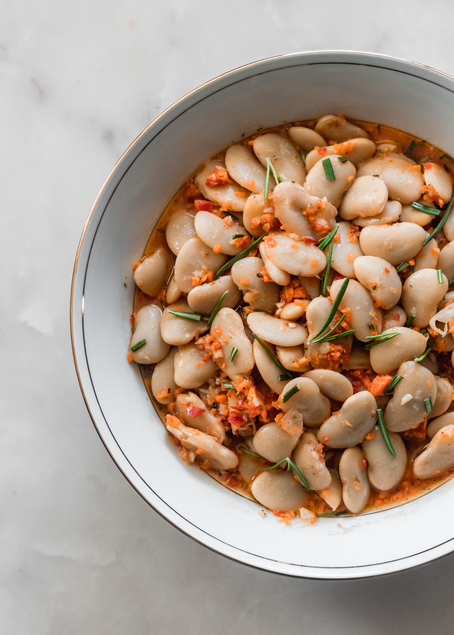 A white bowl filled with marinated white beans, vegetables, and chopped rosemary on a white counter.