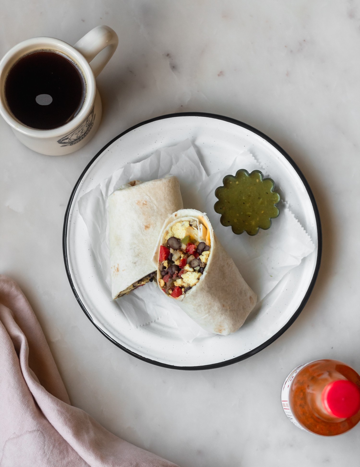 An overhead shot of a white plate with a breakfast burrito on top next to a side of salsa. Next to the burrito is a cup of coffee, hot sauce, and a pink napkin