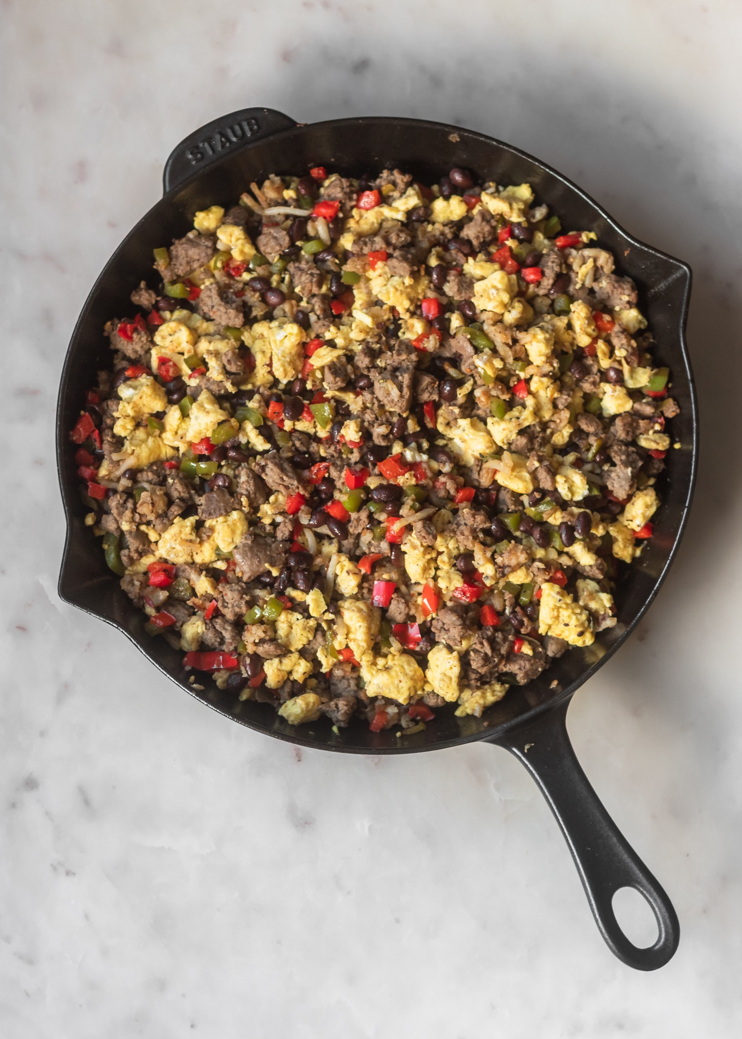 An overhead shot of a black Staub pan filled with eggs, sausage, beans, and vegetables on a light grey counter.