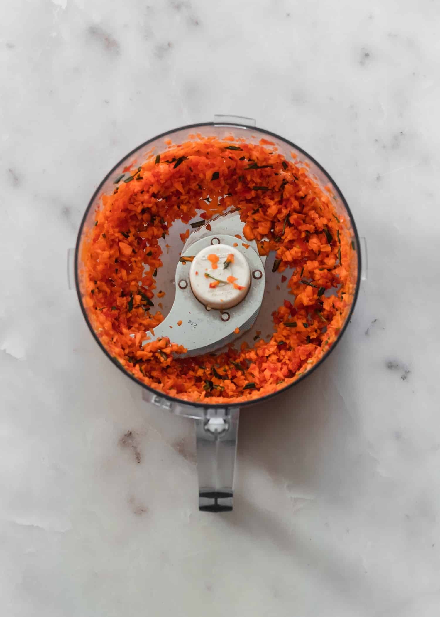 A food processor filled with finely diced vegetables and rosemary, sitting on a white marble counter.