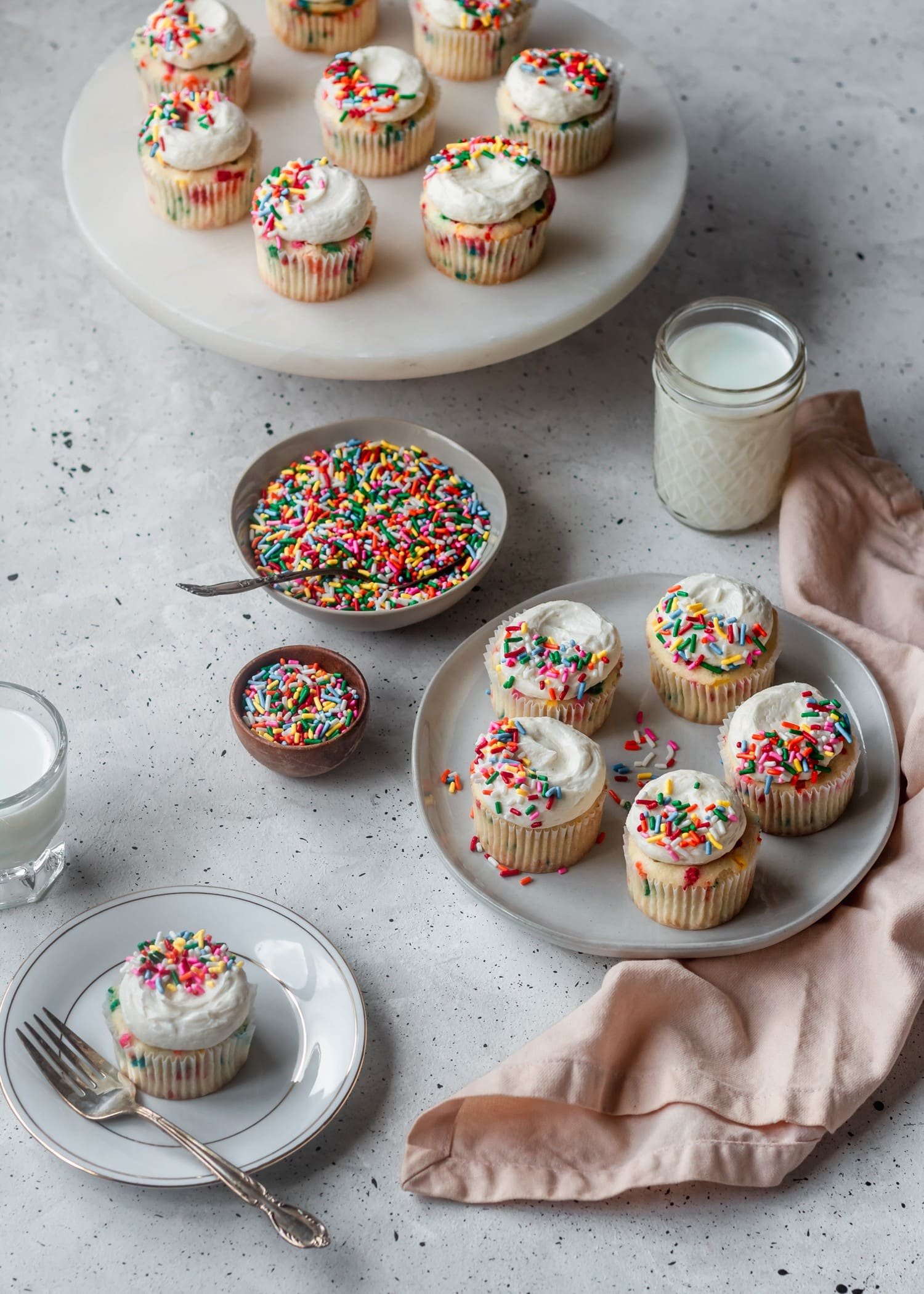 A side shot of a plate of funfetti cupcakes with white frosting and sprinkles on top, surrounded by a cake stand with more cupcakes, a bowl of sprinkles, a pink linen, and glasses of milk.