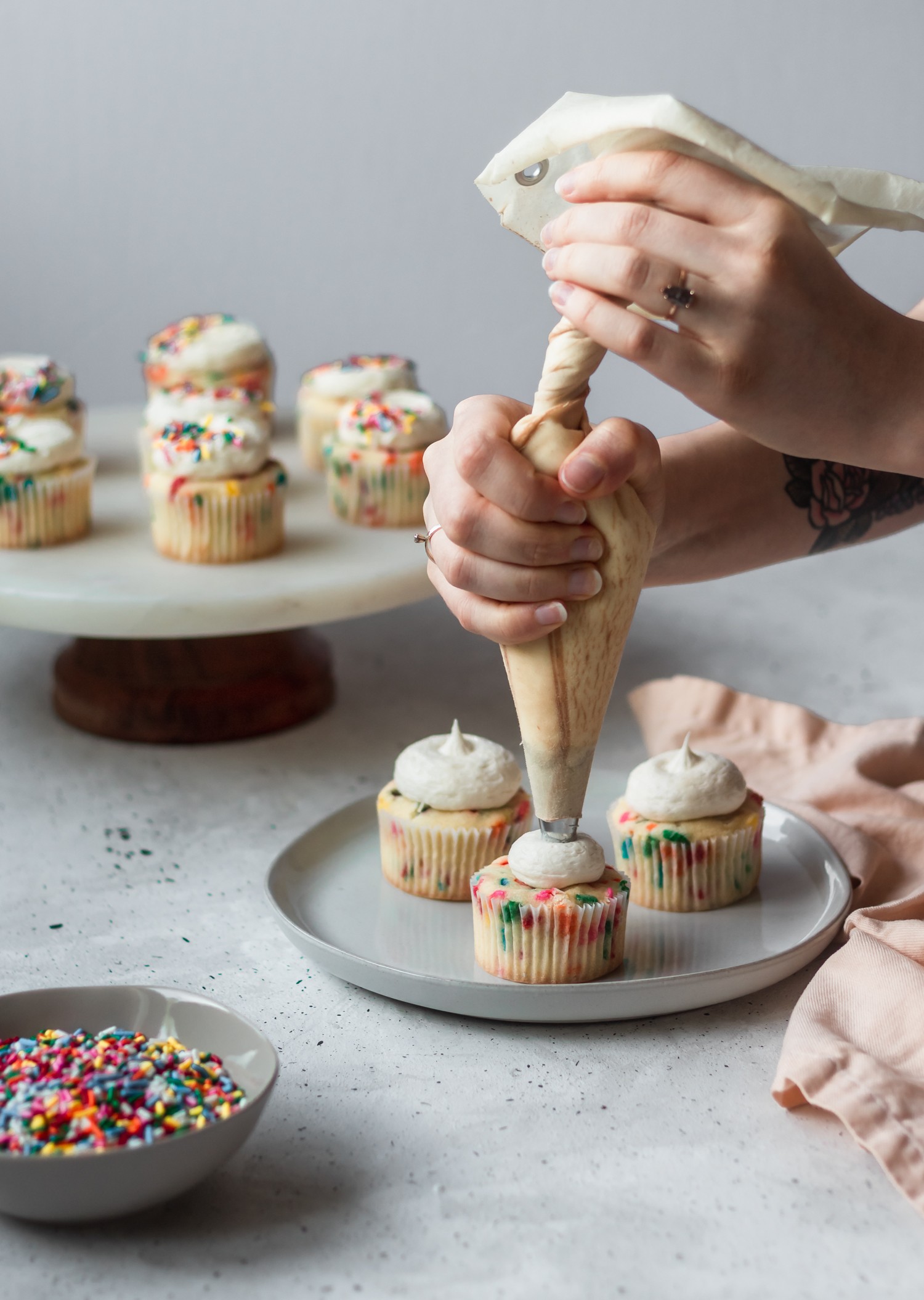 A woman's hands piping frosting onto cupcakes, with a cake stand of cupcakes in the background, and a bowl of sprinkles in the lower left corner with a white and grey background.