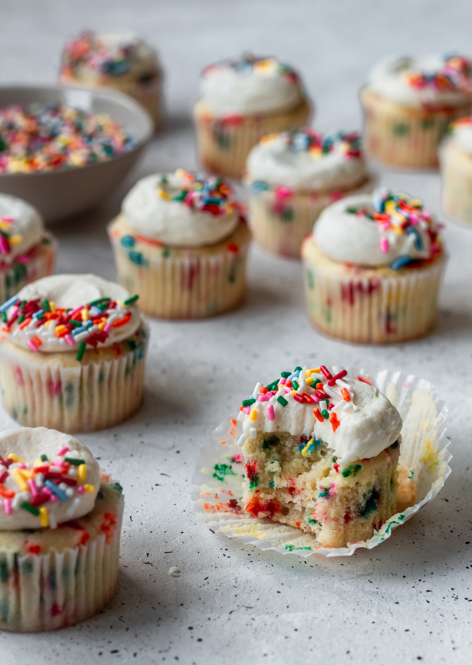 A closeup of a sprinkle cupcake with a bite taken out of it, surrounded by more sprinkle cupcakes on a grey background.