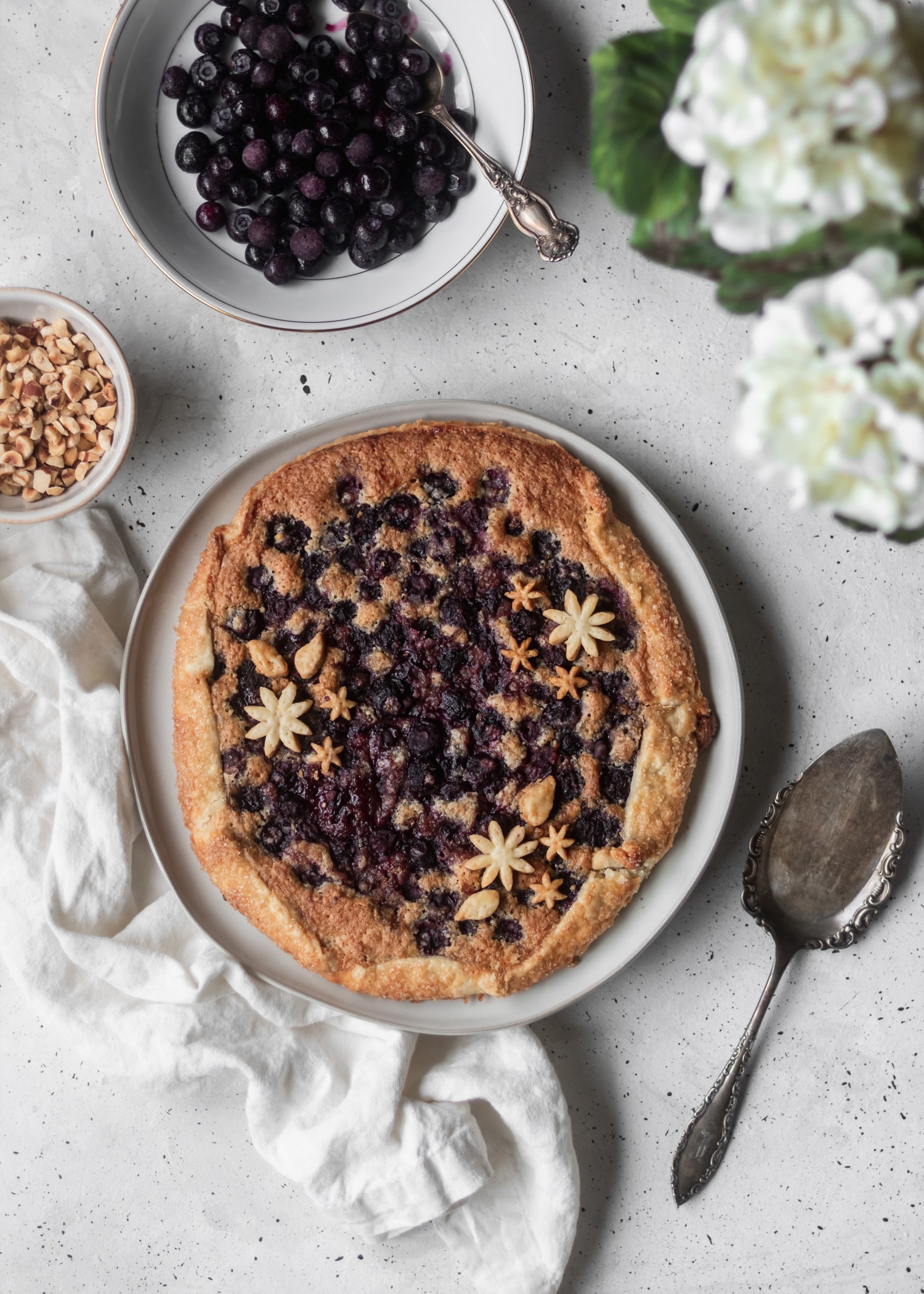A bird's eye view of a blueberry crostata next to a bowl of blueberries, a bowl of hazelnuts, white hydrangeas, and a pie knife on a grey counter.