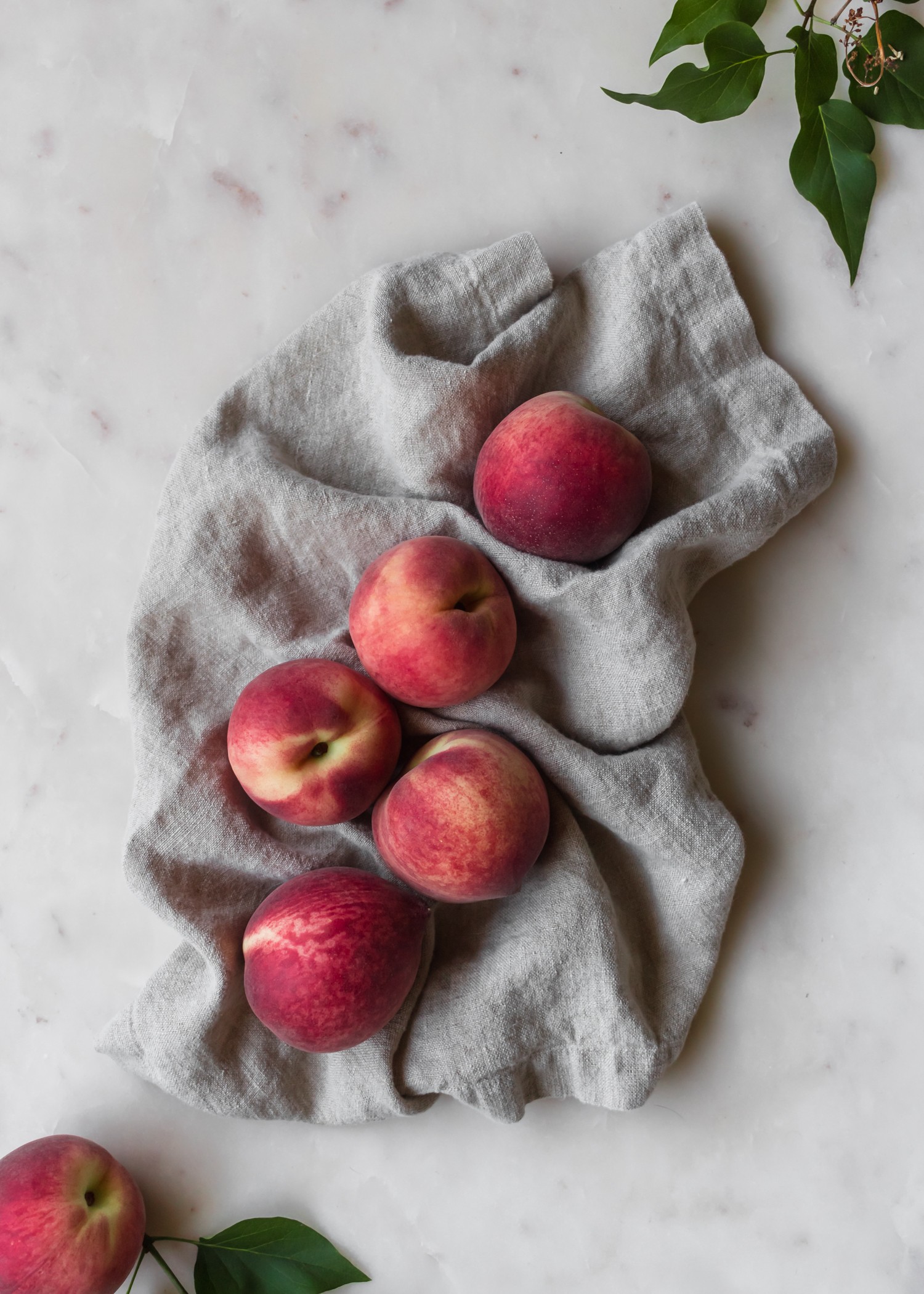 A bird's eye shot of peaches on a beige linen sitting on a marble counter next to green leaves.