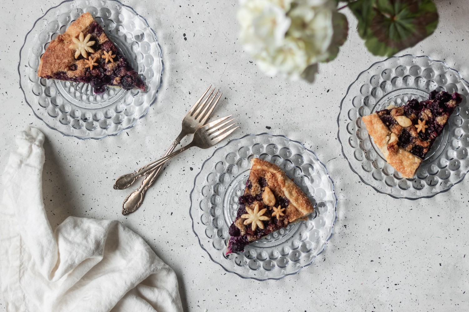 A bird's eye shot of three glass blue plates with slices of berry crostata on top, surrounded by a white linen, forks, and white hydrangeas on a grey table.