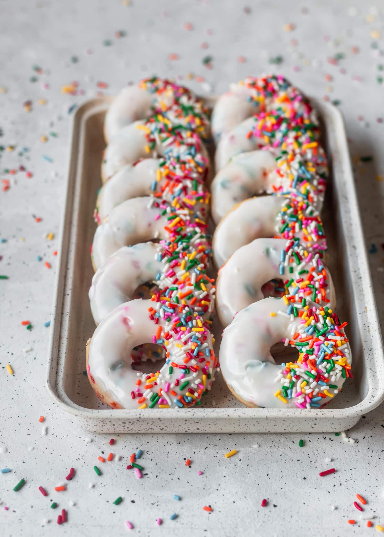 A side shot of a white baking sheet lined with funfetti donuts on a grey table covered in rainbow sprinkles.