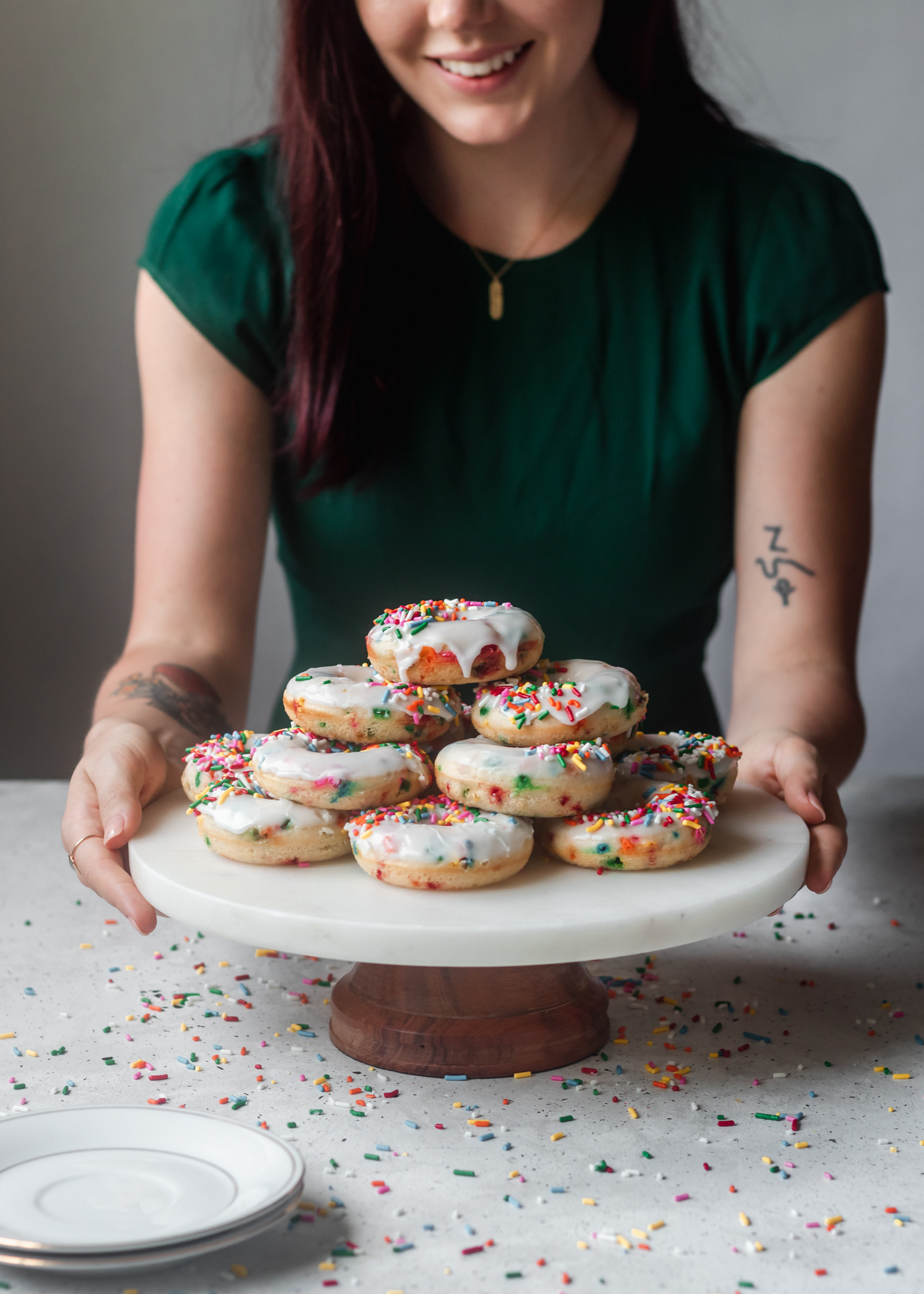 A side shot of a pile of rainbow donuts on a marble cake stand on a white table, with a woman in a dark green dress holding the cake stand.