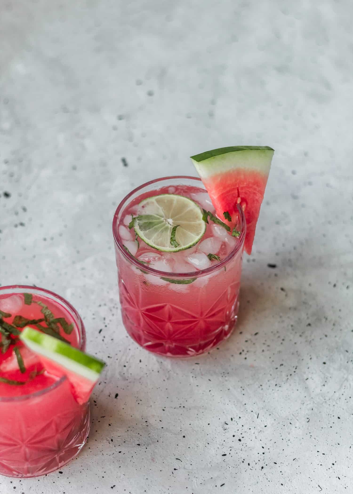 A side shot of a pink cocktail, garnished with mint, melon, and lime on a grey background, next to another cocktail.