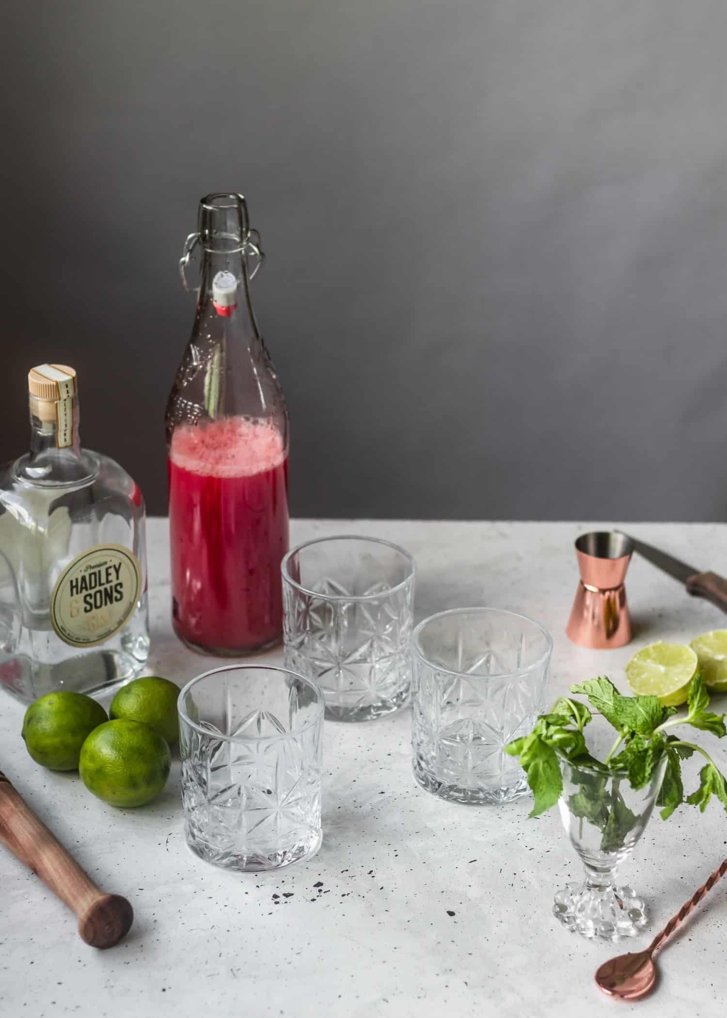 A side shot of old fashioned glasses, surrounded by a bottle of melon juice, an alcohol bottle, limes, mint, and bartending tools on a white table.