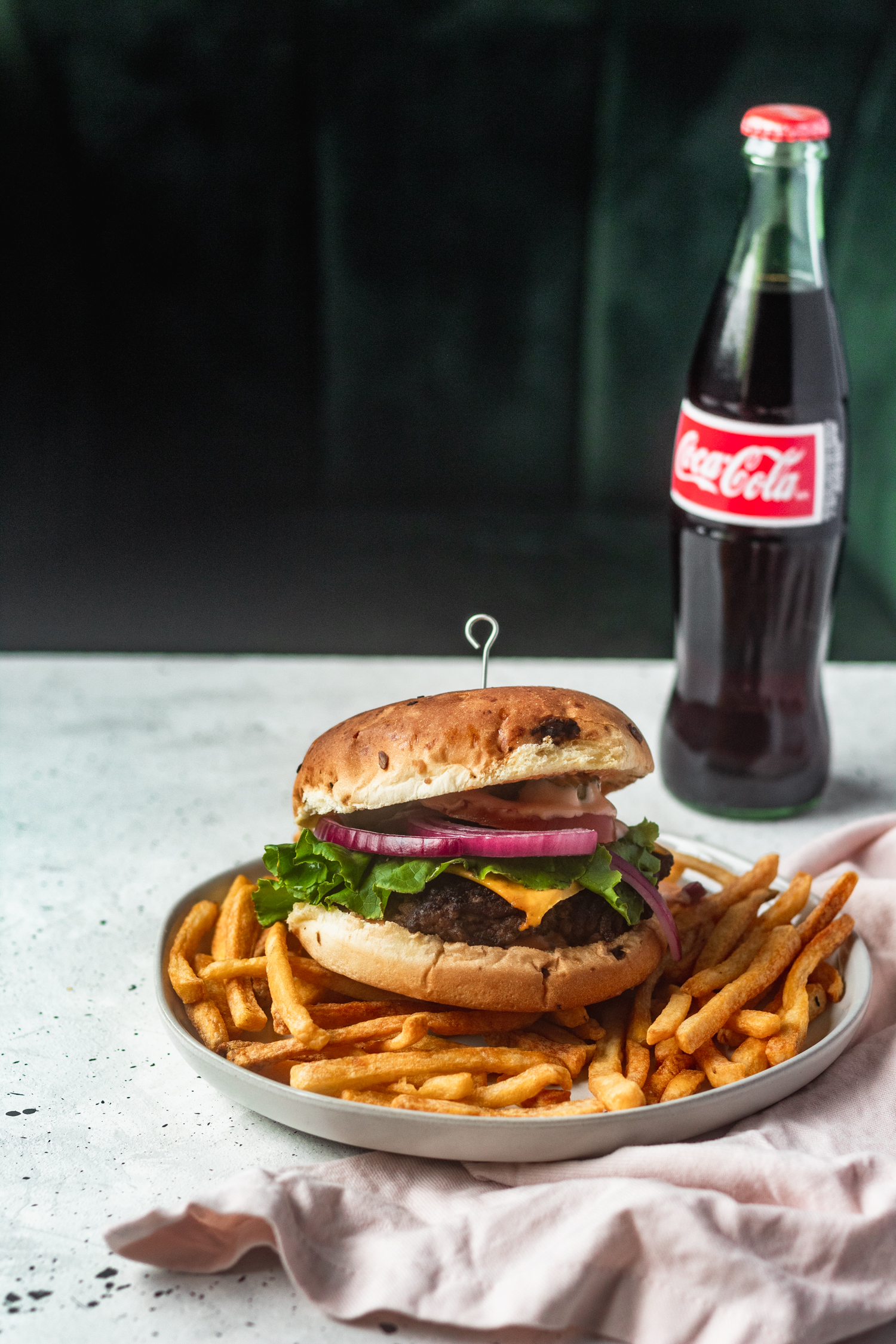 A side shot of a burger placed on top of fries, sitting on a white table with a green background, next to a pink linen and bottle of soda.