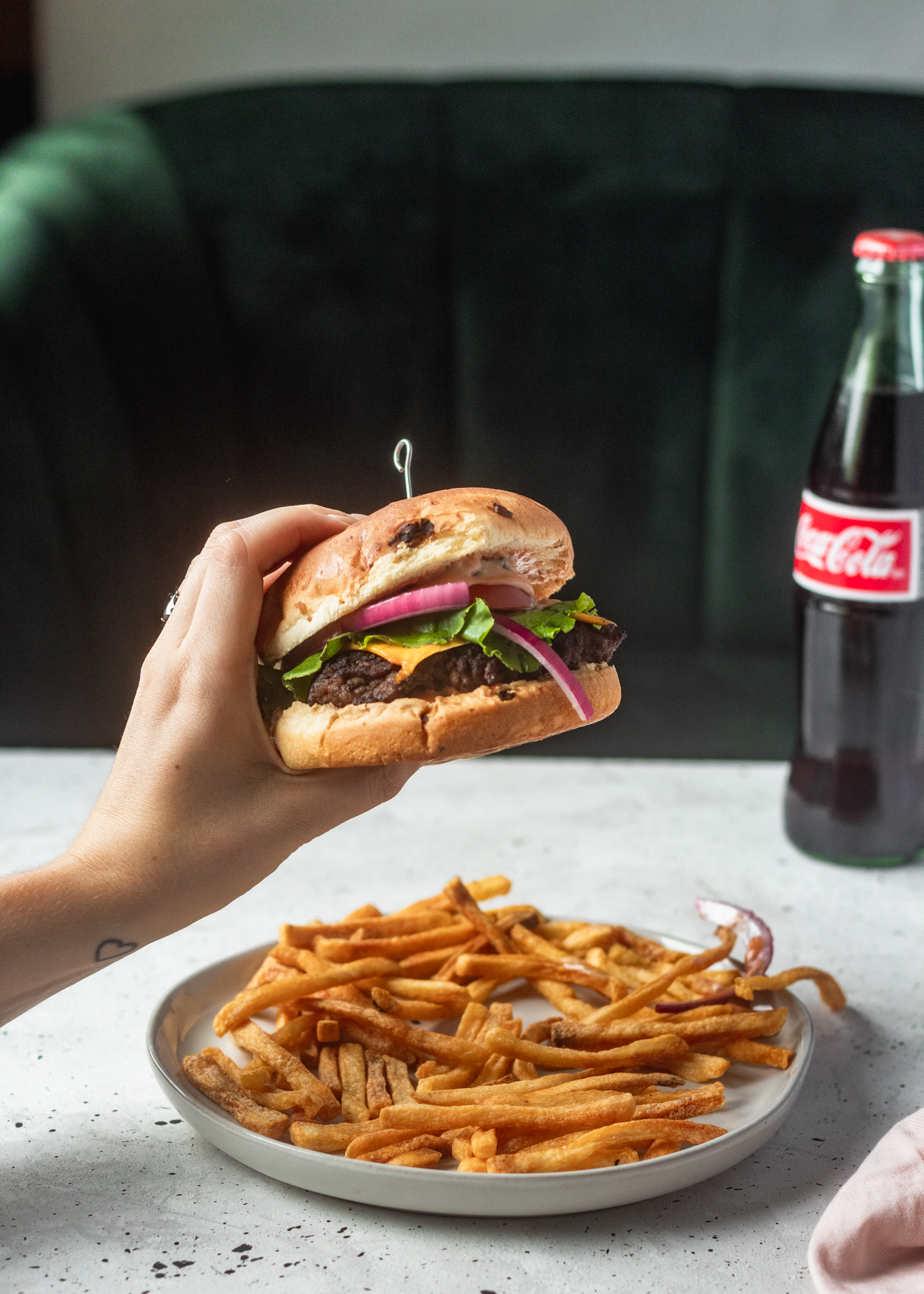 A woman's hand holding a burger, with a white table and green velvet booth in the background. On the table is a plate of fries and a bottle of Coke.