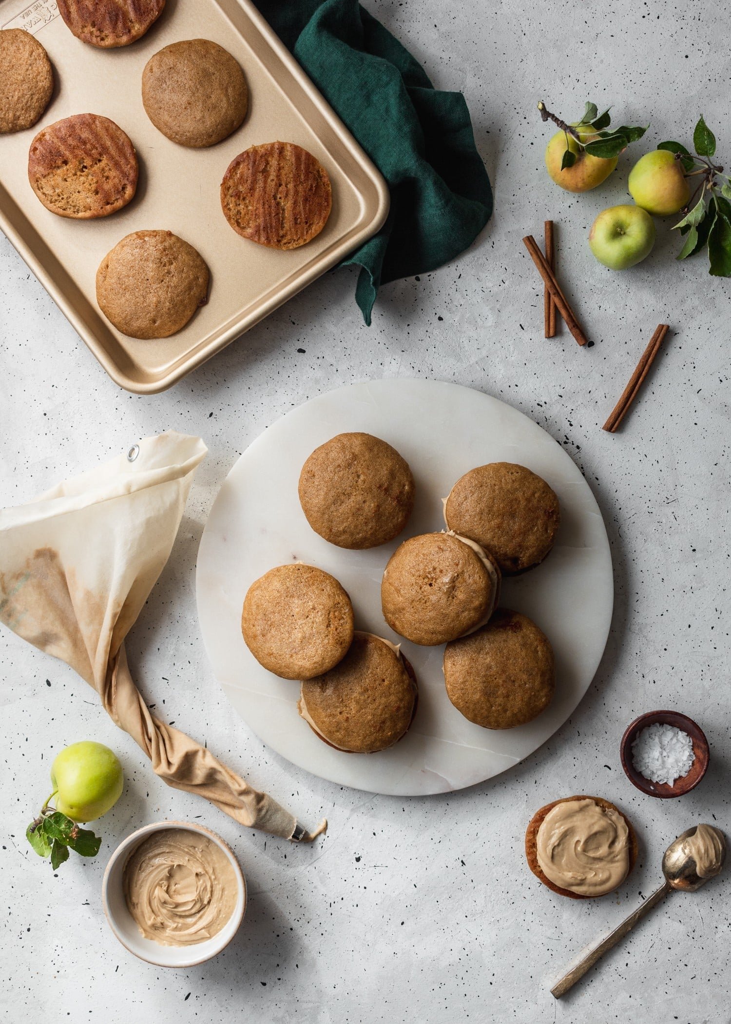 An overhead shot of a white plate filled with apple cider whoopie pies, surrounded by a baking pan, apples, cinnamon, and a piping bag.