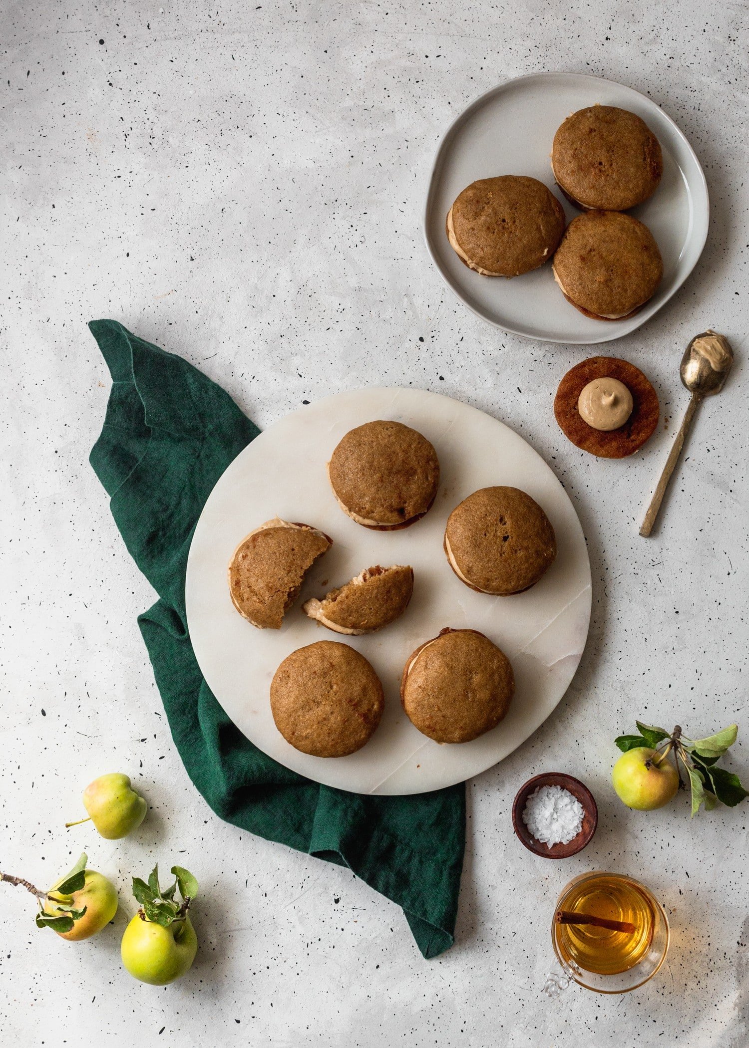 A bird's eye view of a white plate of apple cider whoopie pies on a green linen surrounded by more cookies and apples on a white speckled table.