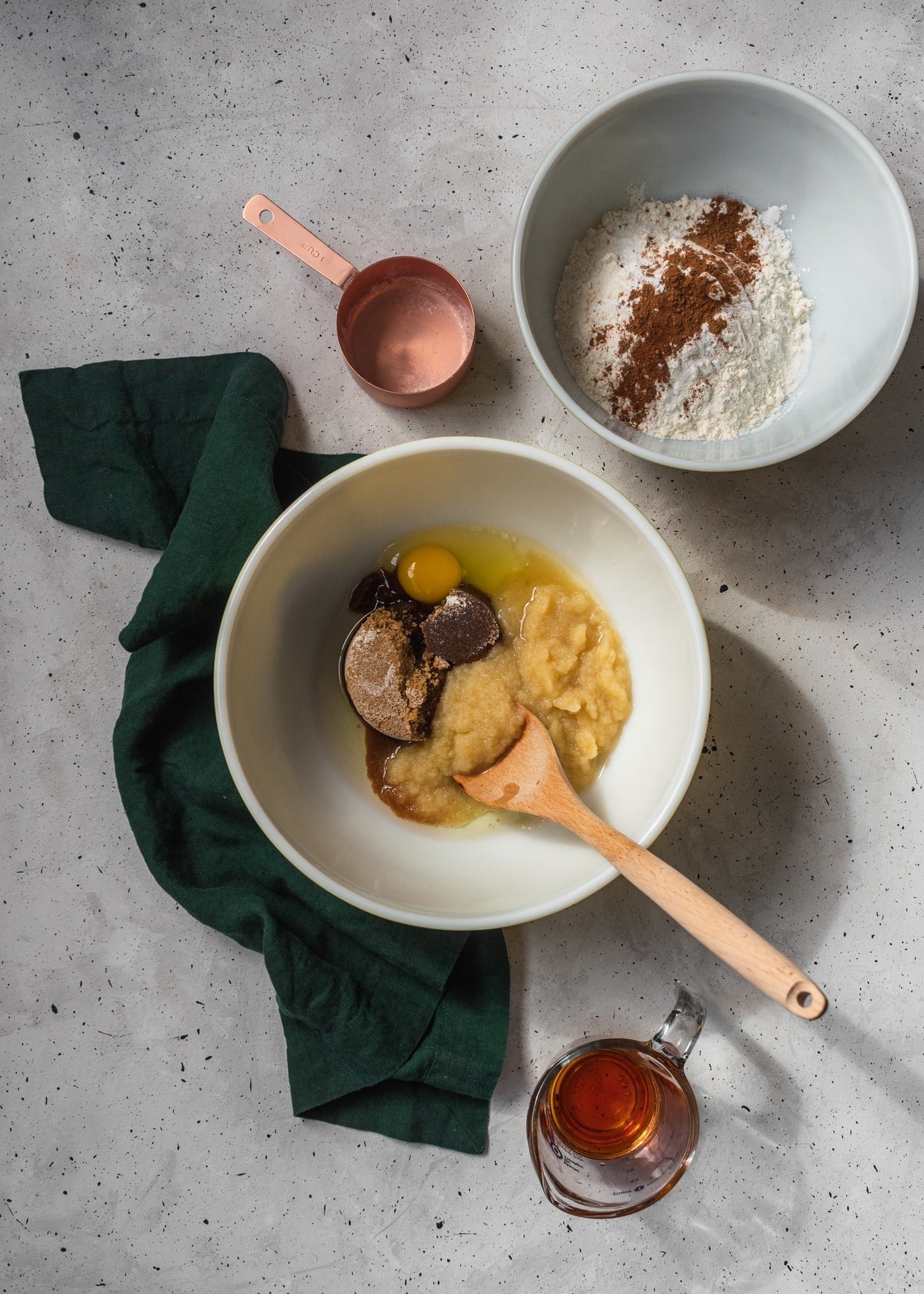 A white mixing bowl with eggs, sugar, applesauce and oil with a wooden spoon sticking out, surrounded by a bowl of flour, a bronze mixing cup, and a green linen on a grey counter.