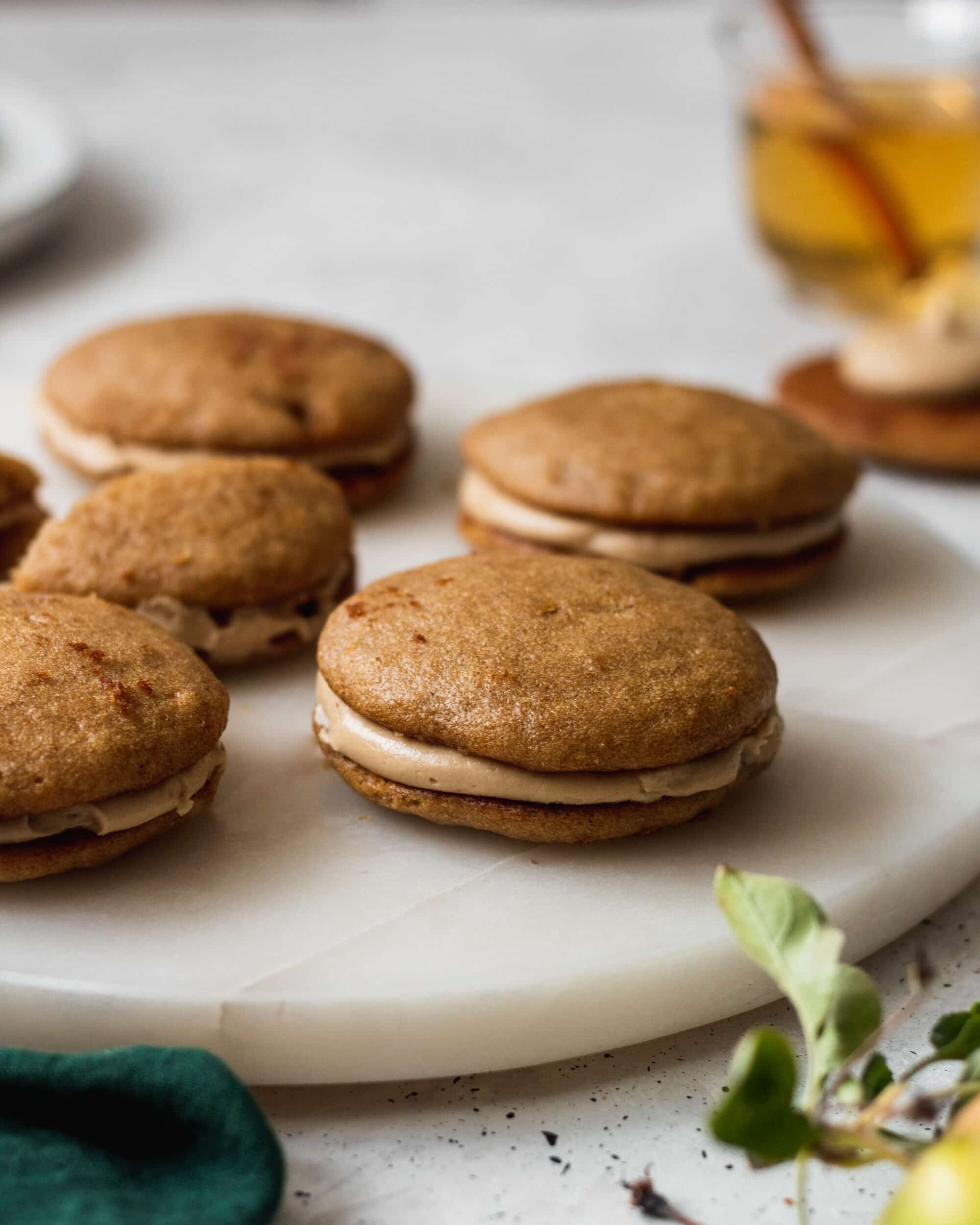 A close-up side shot of apple cider whoopie pies on a marble tray surrounded by an emerald green linen and apples.