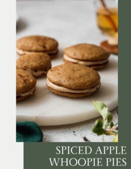 A side image of apple cider whoopie pies with caramel frosting on a white marble plate on a grey table next to a green linen and apple.