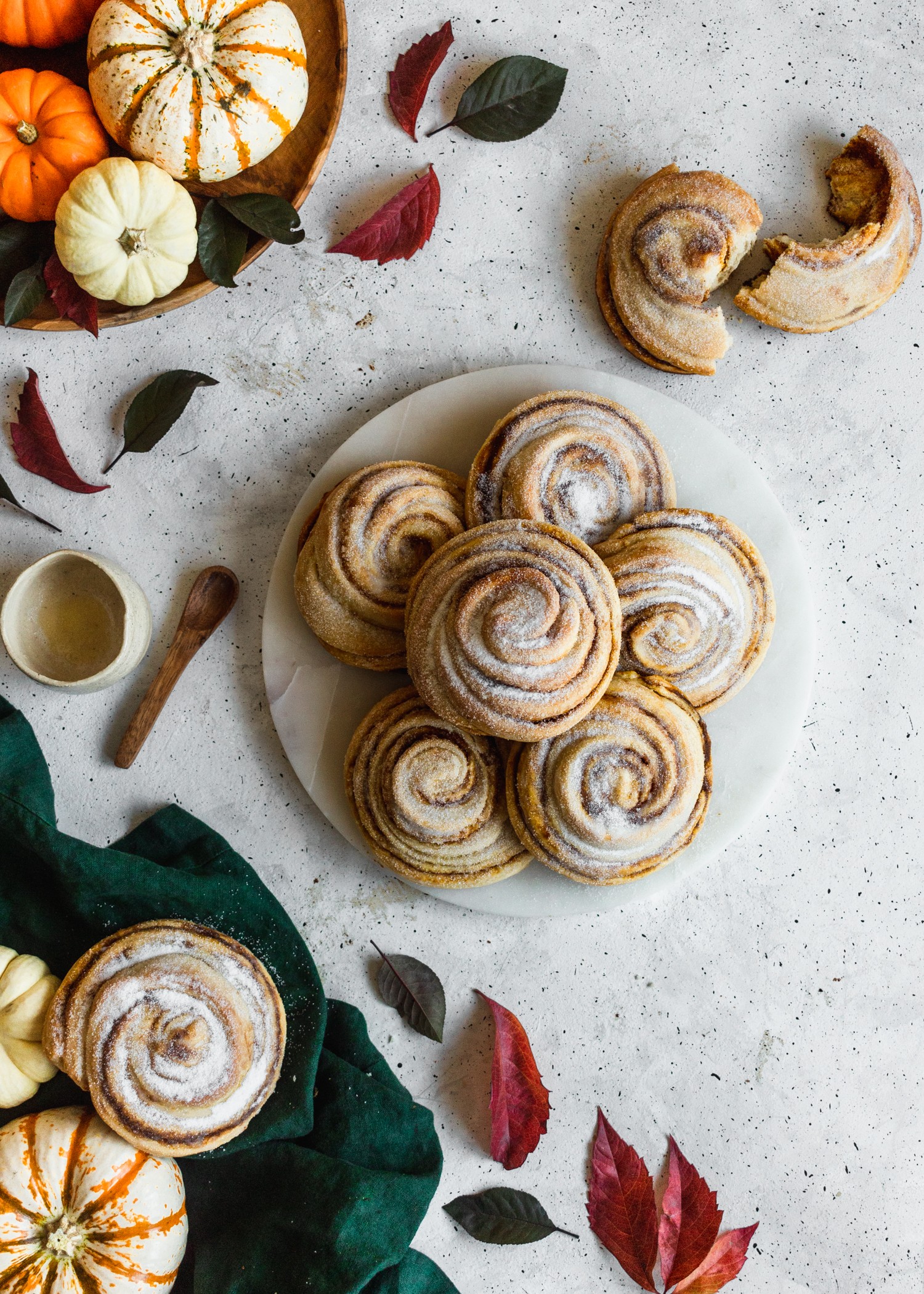 An overhead shot of cinnamon rolls on a white marble plate surrounded by pumpkins, leaves, and a dark green linen on a grey background.