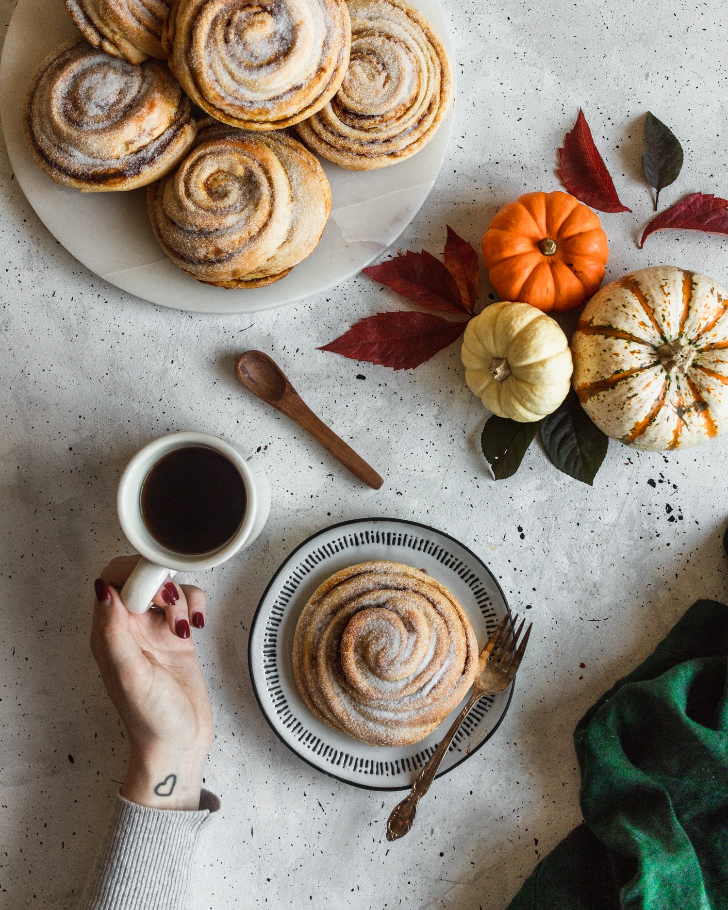 A bird's eye view photo of a pumpkin morning bun on a black and white plate surrounded by a green napkin, pumpkins, and a woman's hand holding a cup of coffee.