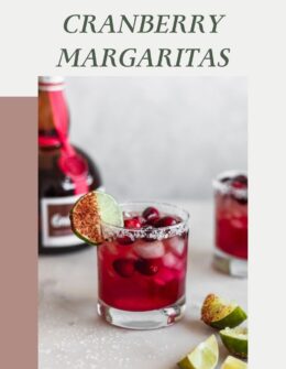 A side image of a cranberry margarita garnished with a lime wedge on a white marble counter with another margarita and a bottle of Grand Marnier in the background.