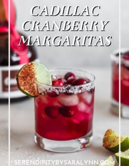 A side image of a cranberry margarita garnished with a lime wedge on a white marble counter with another margarita and a bottle of Grand Marnier in the background.