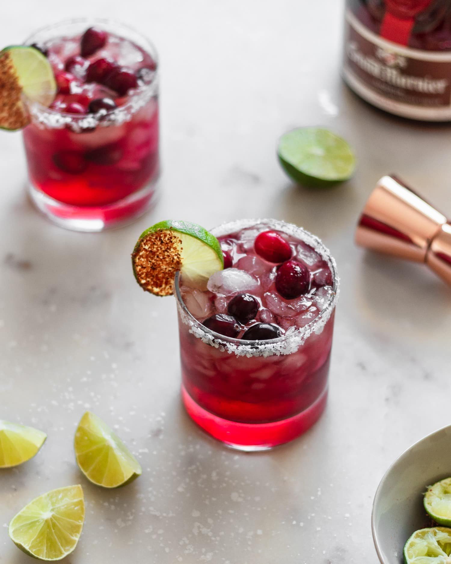 Two Cadillac cranberry margaritas on a marble counter next to lime wedges and a bottle of Grand Marnier.