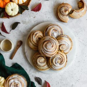 An overhead photo of cinnamon swirl buns with pumpkin filling on a white plate surrounded by a green linen and pumpkins on a grey background.
