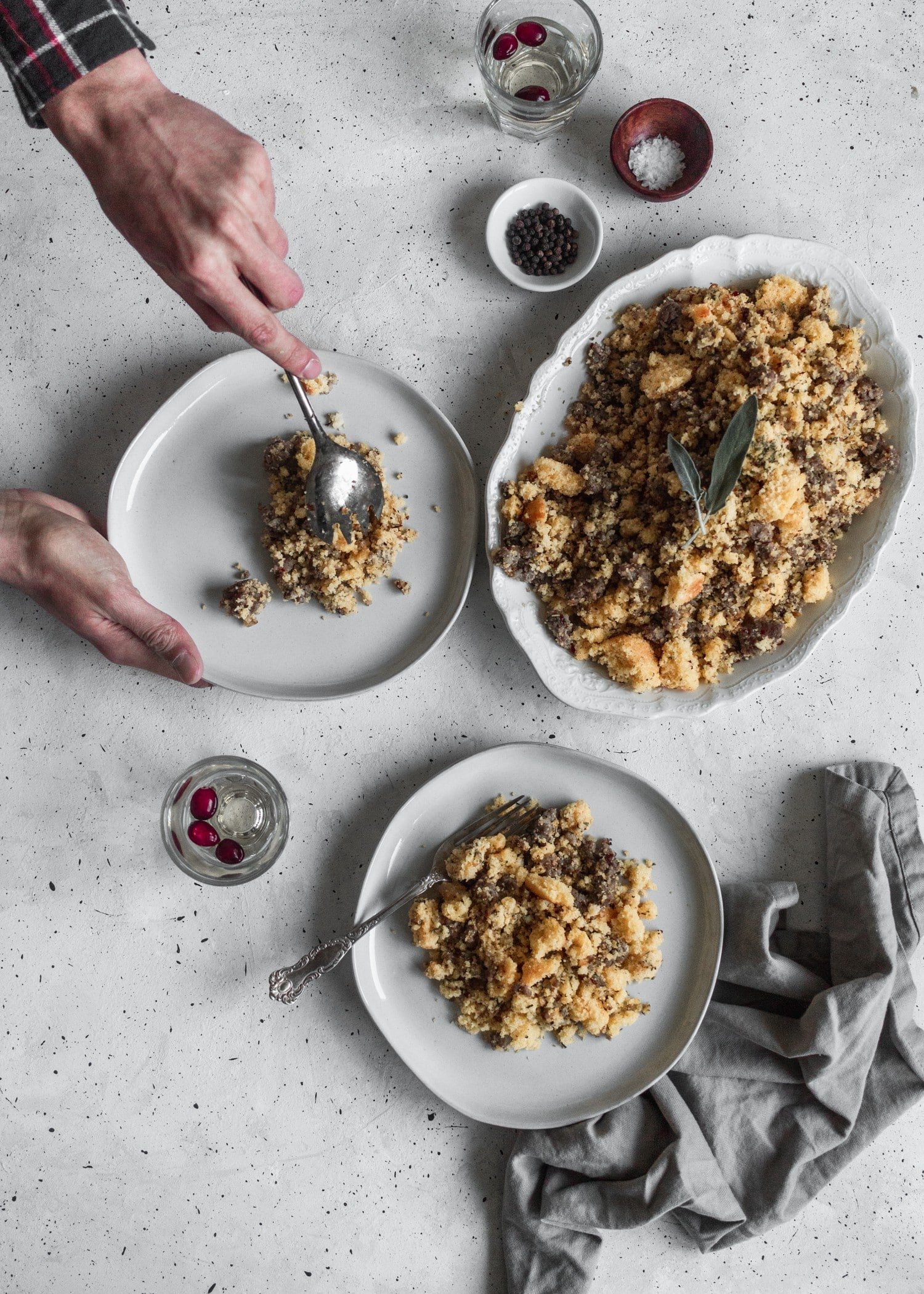 A bird's eye view shot of a man's hand reaching for a plate of Thanksgiving dressing next to plates of dressing and wine on a grey table.