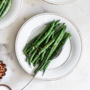 An overhead photo of green beans on a white plate surrounded by more plates on a white marble table.