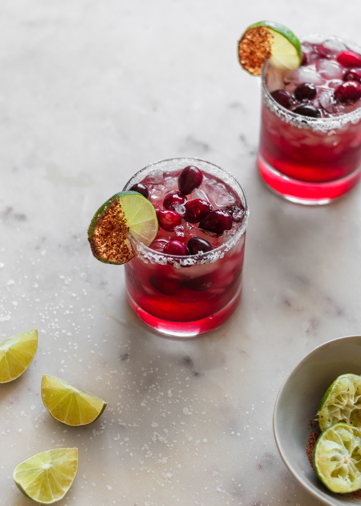 A 45 degree shot of two cranberry margaritas on a white counter surrounded by limes and salt.