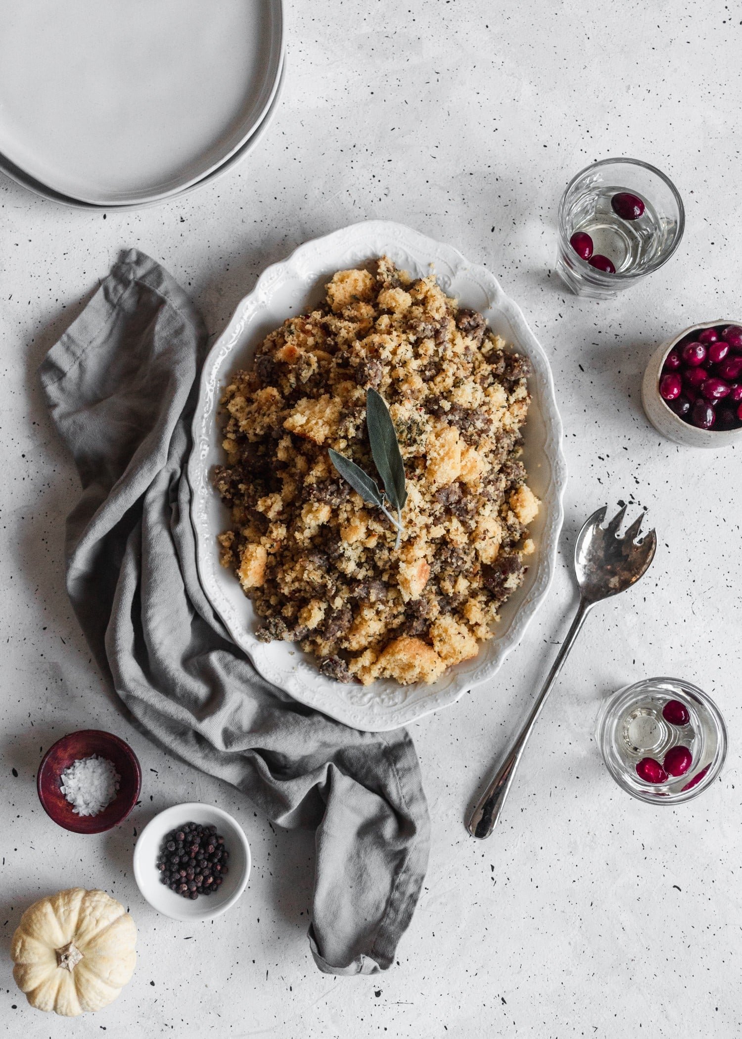 An overhead photo of cornbread stuffing on an oval white plate, set on a grey table surrounded by glasses of white wine, a bowl of cranberries, salt, pepper, and a grey linen.