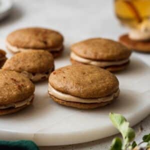 A side photo of apple cider whoopie pies on a marble plate surrounded by apples and a green linen.