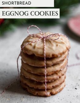 A closeup side image of a stack of eggnog cookies tied with striped red and white twine on a white speckled table. In the background is a vintage red plate and pine branch.