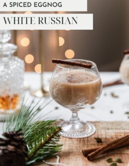 A side image of an eggnog white Russian cocktail on a wood board next to a pine branch, bottle of liquor, and spices on a white counter. In the background is a grey wall covered in holiday lights.