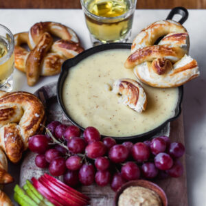 A side photo of cheese fondue surrounded by pretzels, beer, and fruit.