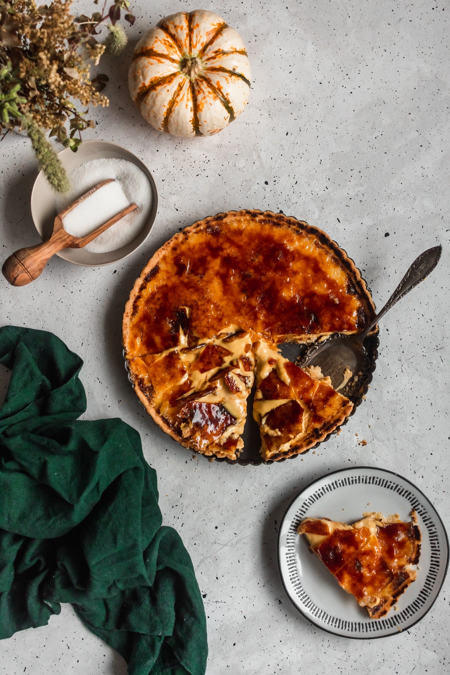 An overhead photo of a sliced creme brulee tart on a grey background surrounded by a green linen, grey plate, and a bowl of sugar.