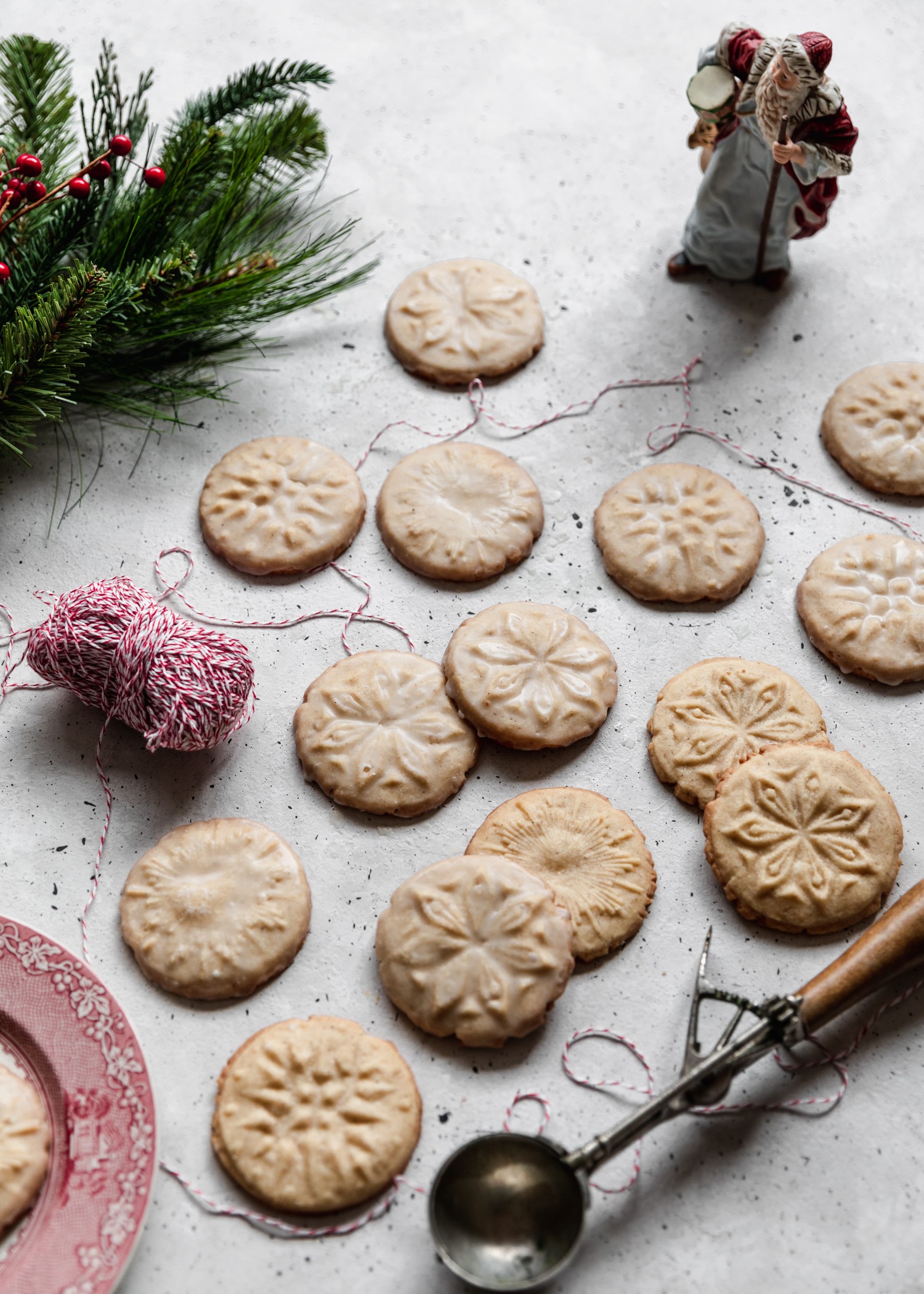 A side image of eggnog cookies surrounded by pine branches, a Santa figurine, and baking twine on a grey counter.