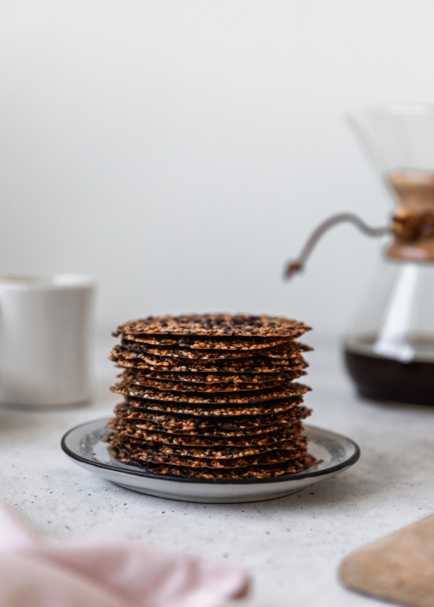 A side image of a stack of sesame lace cookies with a Chemex and coffee cup in the background on a white table.