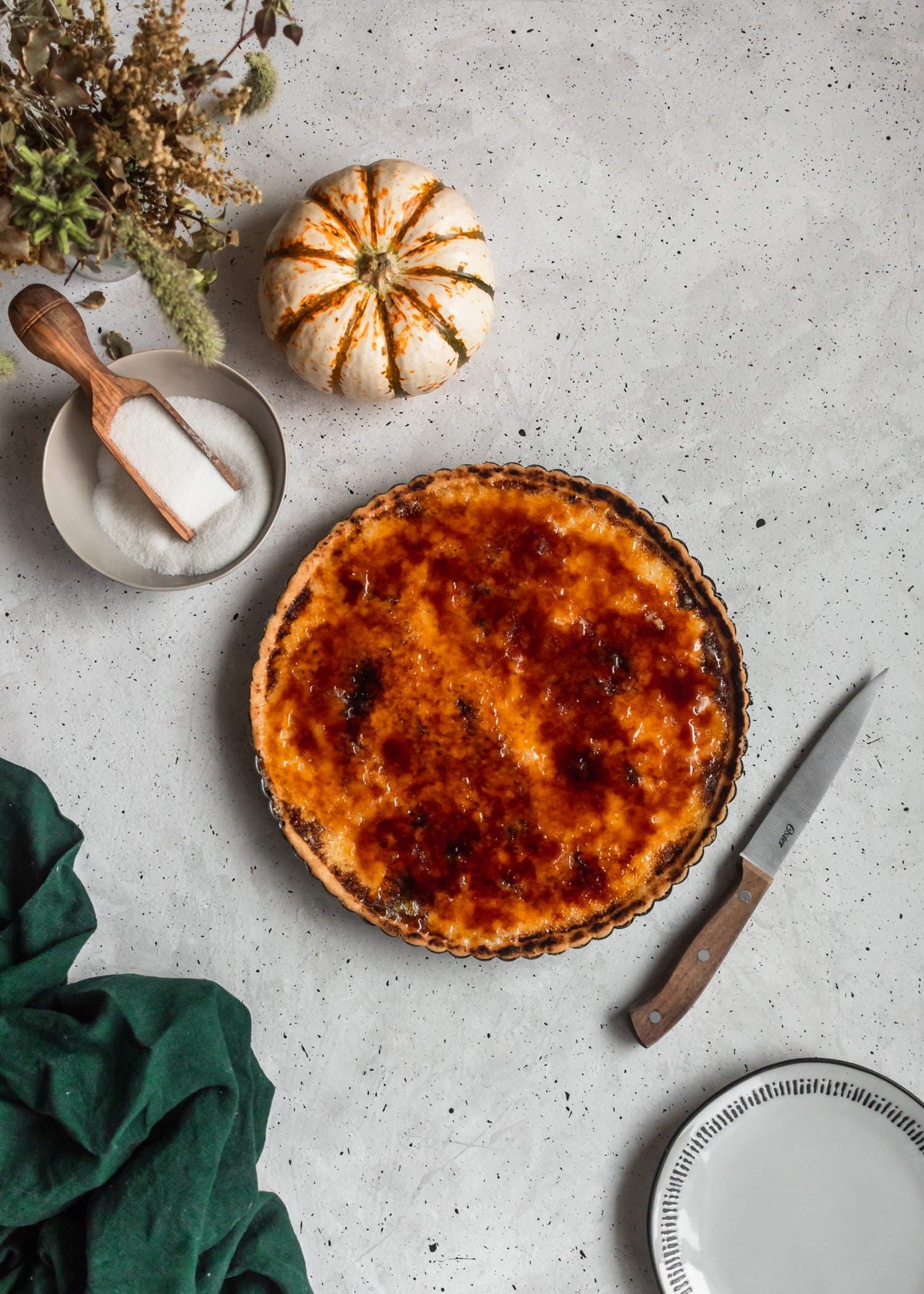 A bird's eye photo of a creme brulee tart on a grey table placed next to a green towel, wood knife, and grey ceramics.