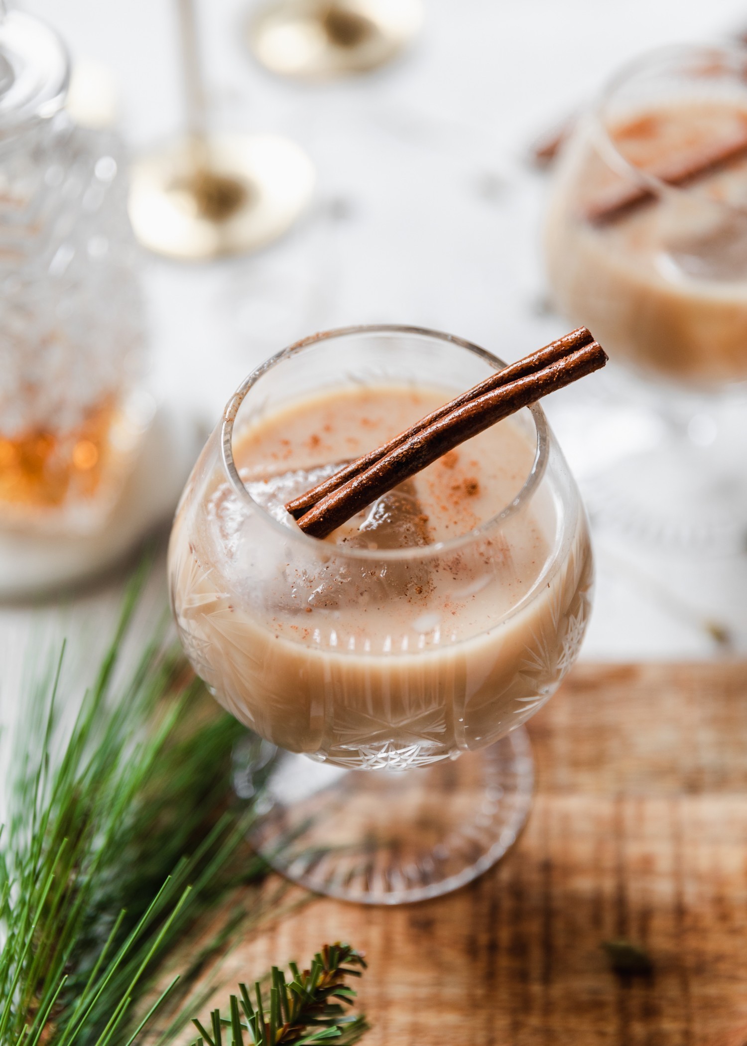 A 45 degree photo of eggnog White Russians on a wood platter placed on a white table with alcohol and pine in the background.