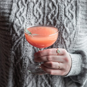 A side image of a woman wearing a grey sweater holding a pink cocktail.