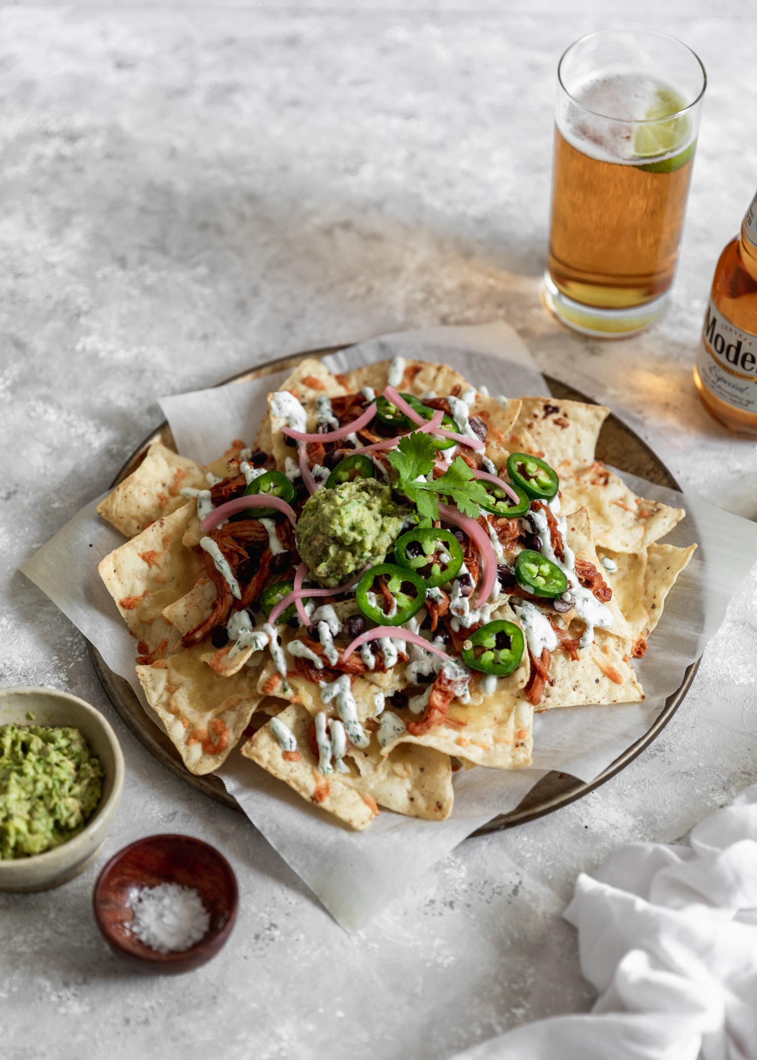 A side image of a plate of BBQ chicken nachos on a grey counter surrounded by beer, a bowl of guacamole, and a wooden bowl of salt.