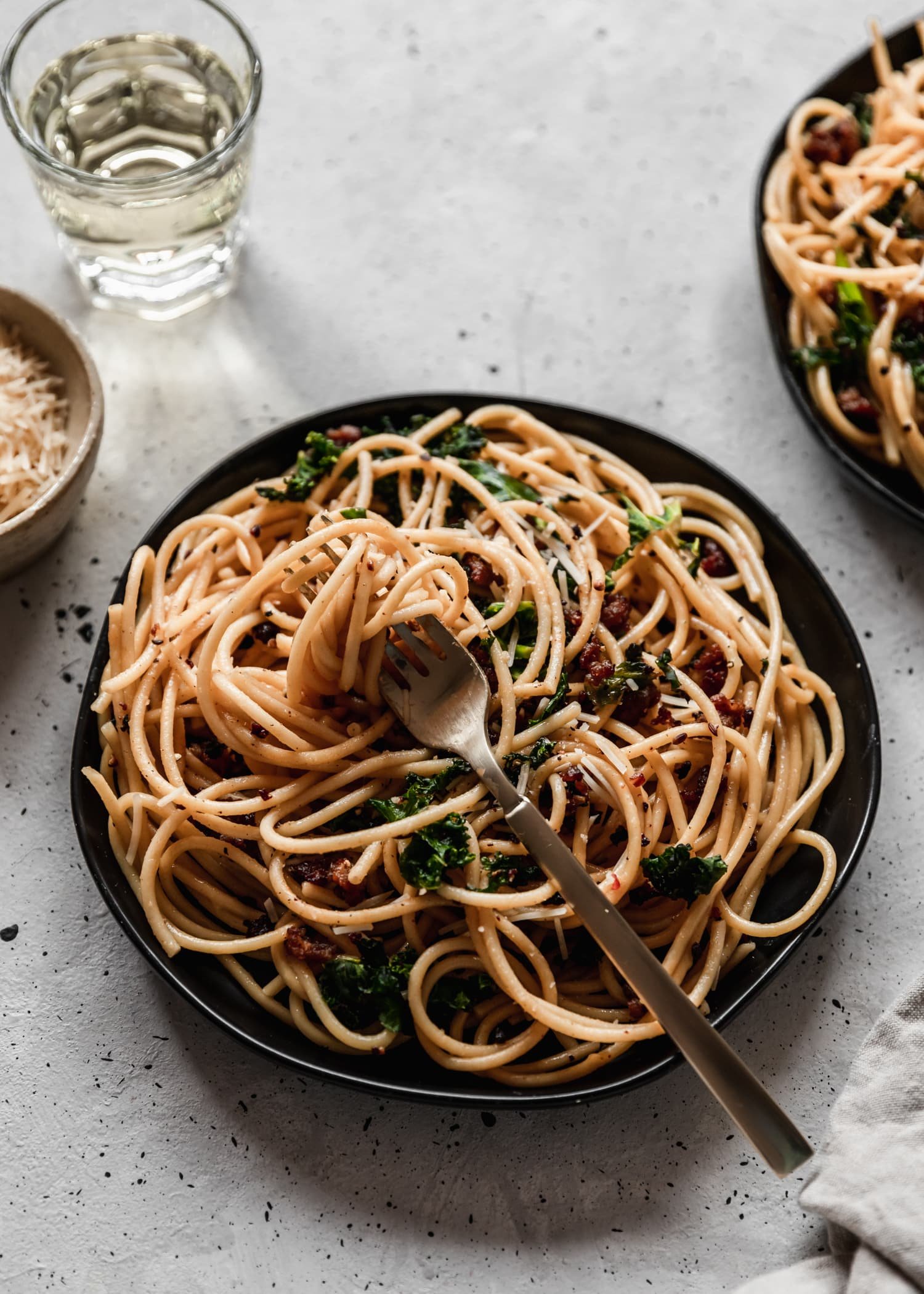 A closeup side image of a gold fork twisted with pasta on a black plateplaced on a grey counter surrounded by more pasta, a bowl of Parm, and a glass of white wine.