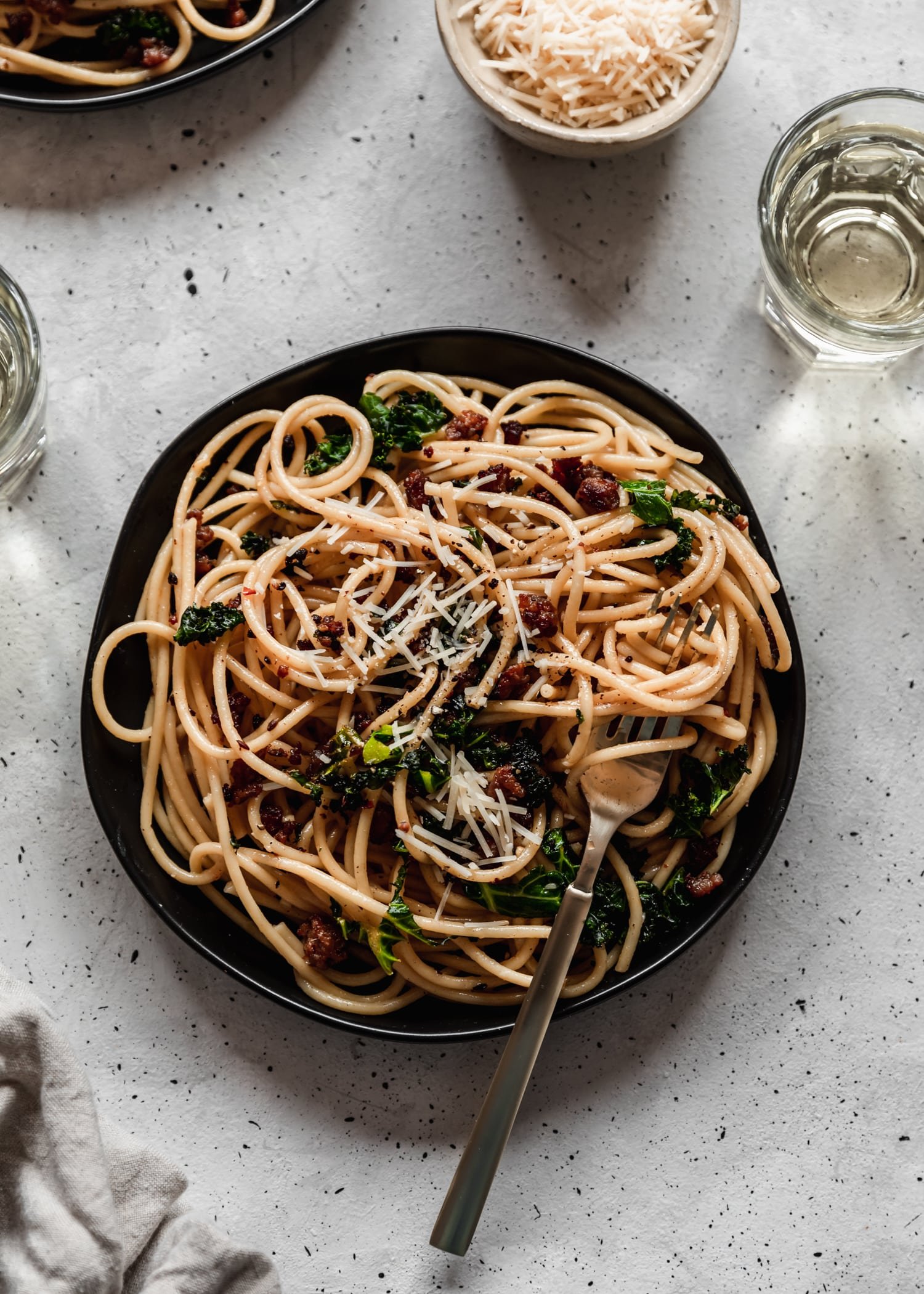 A closeup overhead of a gold fork twirled with bucatini carbonara with sausage and greens on a black plate placed on a grey table surrounded by glasses of white wine, a bowl of parm, and a grey linen.