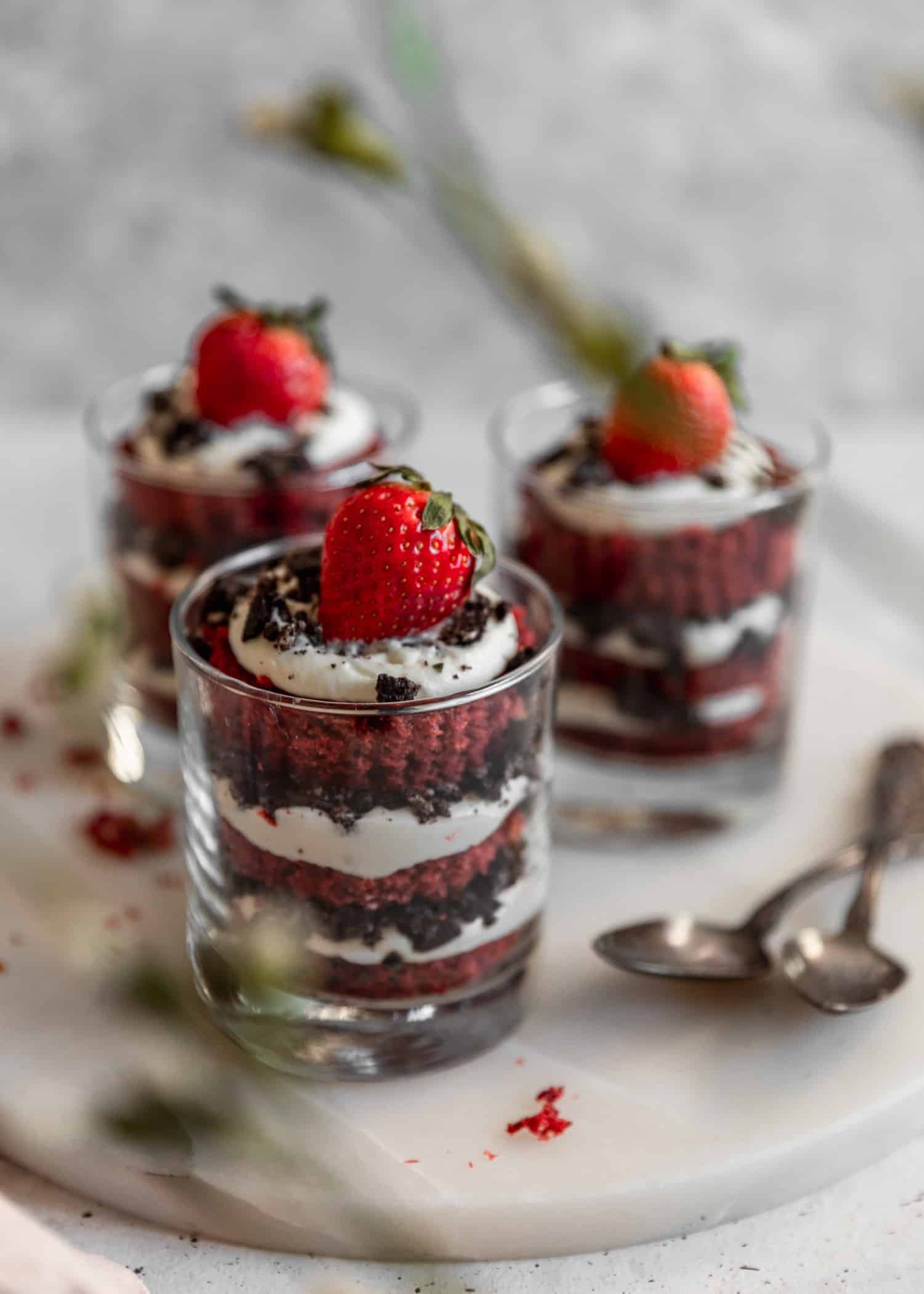 Three layered puddings with red cake, white cream, Oreos, and strawberries on a marble trey with a grey background and thin green and white flower buds blurred in the front.