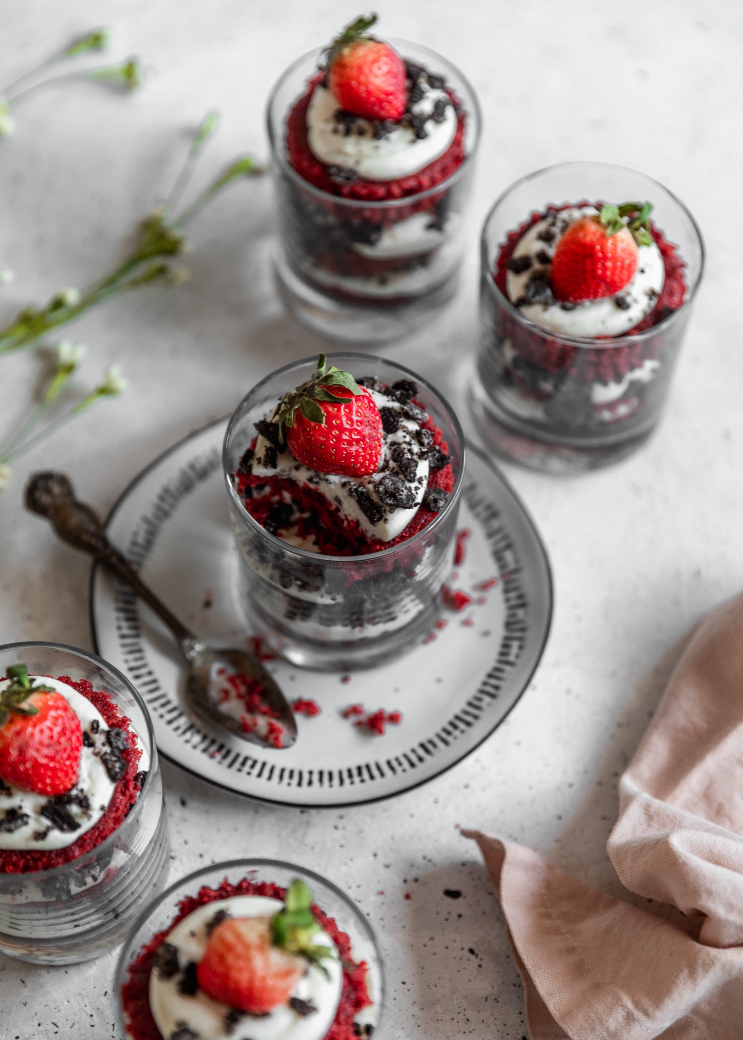 Five cheesecake red velvet trifles on a grey table in a curved shape. The middle trifle is on a black and white plate with a spoon, and there is a pink linen in the right lower corner.