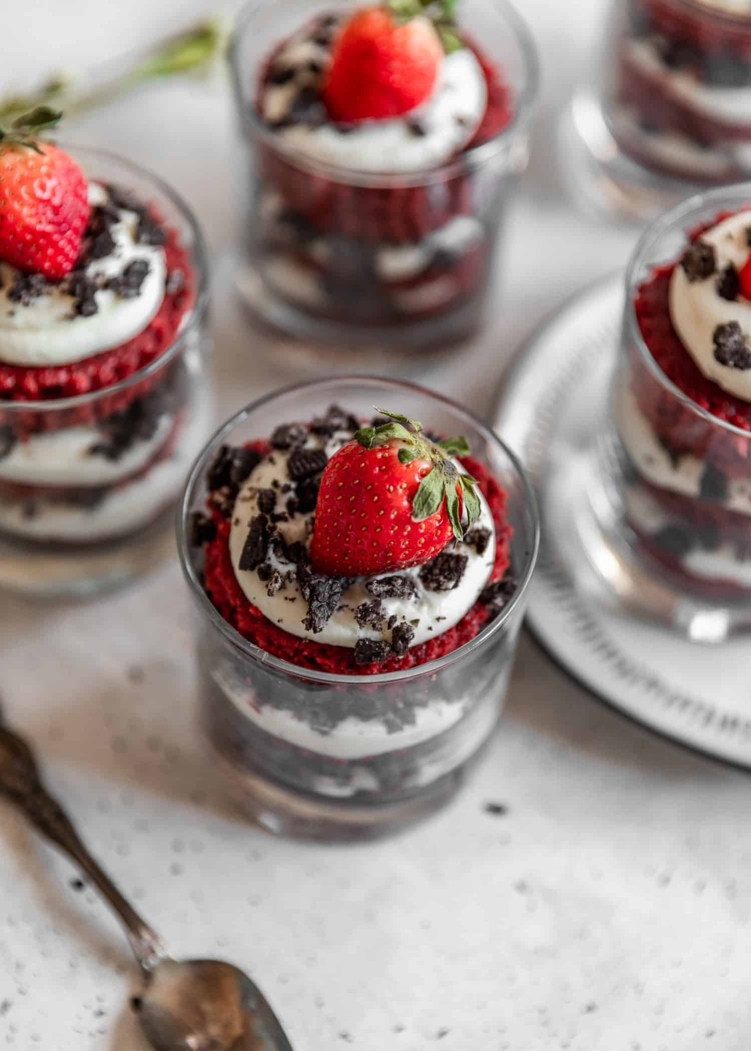 A 45 degree shot two red velvet trifles with cheesecake filling, crushed chocolate cookies, and a strawberry on a grey table surrounded by three more trifles and a spoon.